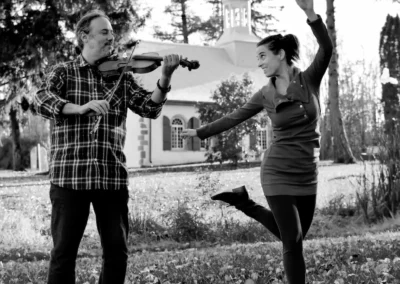 Photo noir et blan Gabriel Girouard joue du violon pendant que Mélissandre Tremblay-Bourassa danse la gigue devant une petite église blanche.