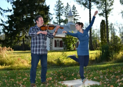 Gabriel Girouard joue du violon pendant que Mélissandre Tremblay-Bourassa danse la gigue devant une petite église blanche.