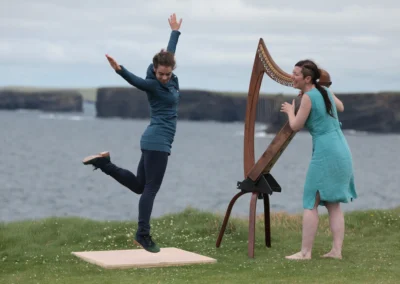 Danseuse de gigue en plein mouvement devant une harpiste irlandaise, avec les falaises et l'océan en arrière-plan.