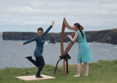 Danseuse de gigue en plein mouvement devant une harpiste irlandaise, avec les falaises et l'océan en arrière-plan.