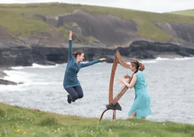 Mélissandre Tremblay-Bourassa saute en giguant devant Úna Ní Fhlannagáin jouant de la harpe sur une falaise en Irlande.