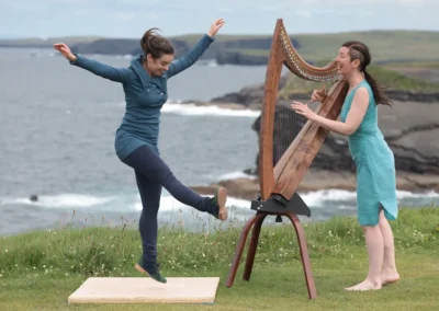 Danseuse de gigue en pleine extension devant une harpiste irlandaise, avec les falaises et l'océan en arrière-plan.